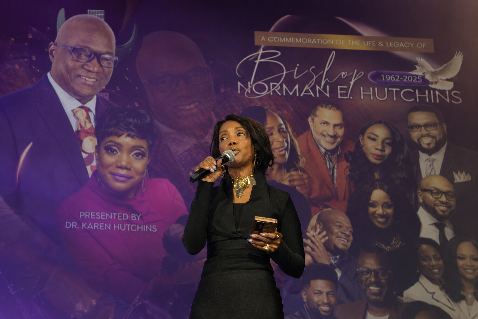 Dr. Karen Hutchins speaking into a microphone on stage in front of a large purple tribute backdrop commemorating the life and legacy of Bishop Norman E. Hutchins